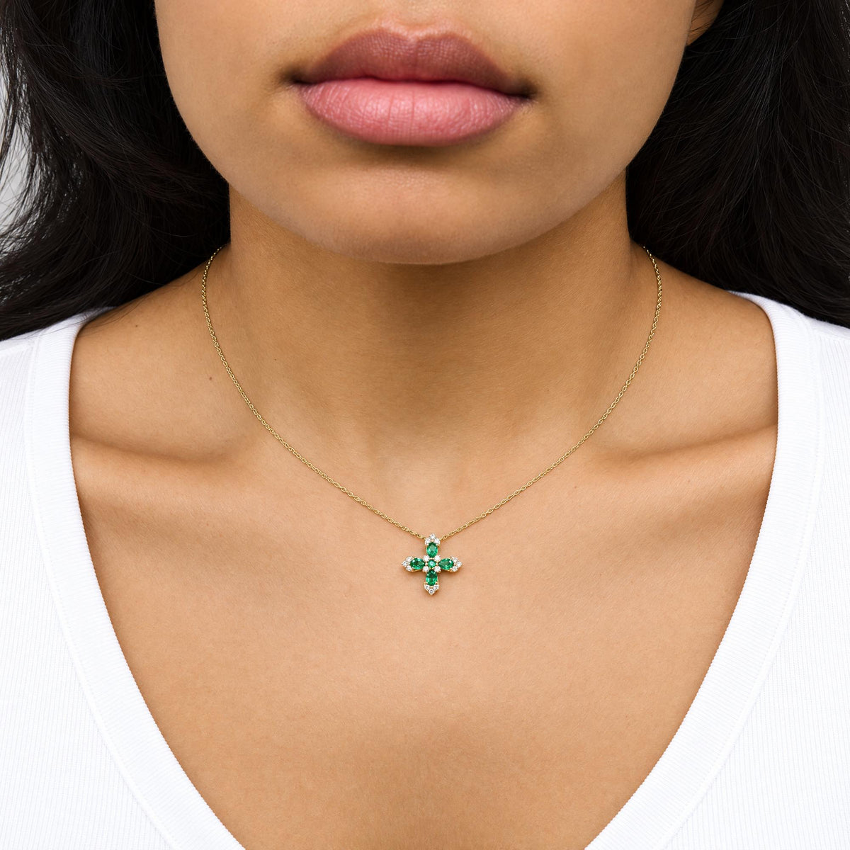 Close-up front view of a model wearing a yellow metal necklace with a cross-shaped pendant featuring pear and round green gemstones surrounded by round white gemstones in prong settings.