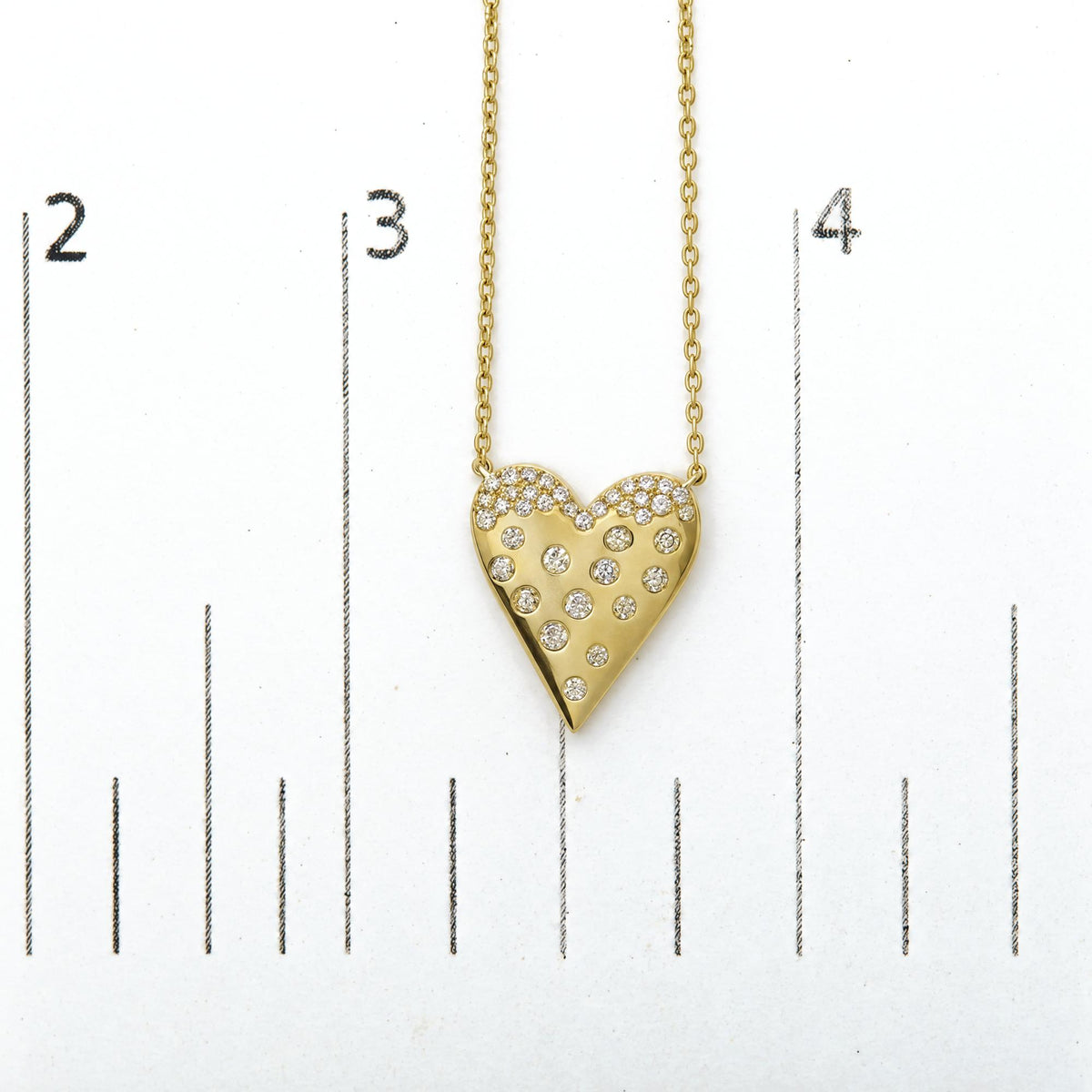 Close-up front view of a yellow metal heart-shaped necklace pendant adorned with multiple white round gemstones in pave setting, shown on a measurement scale.