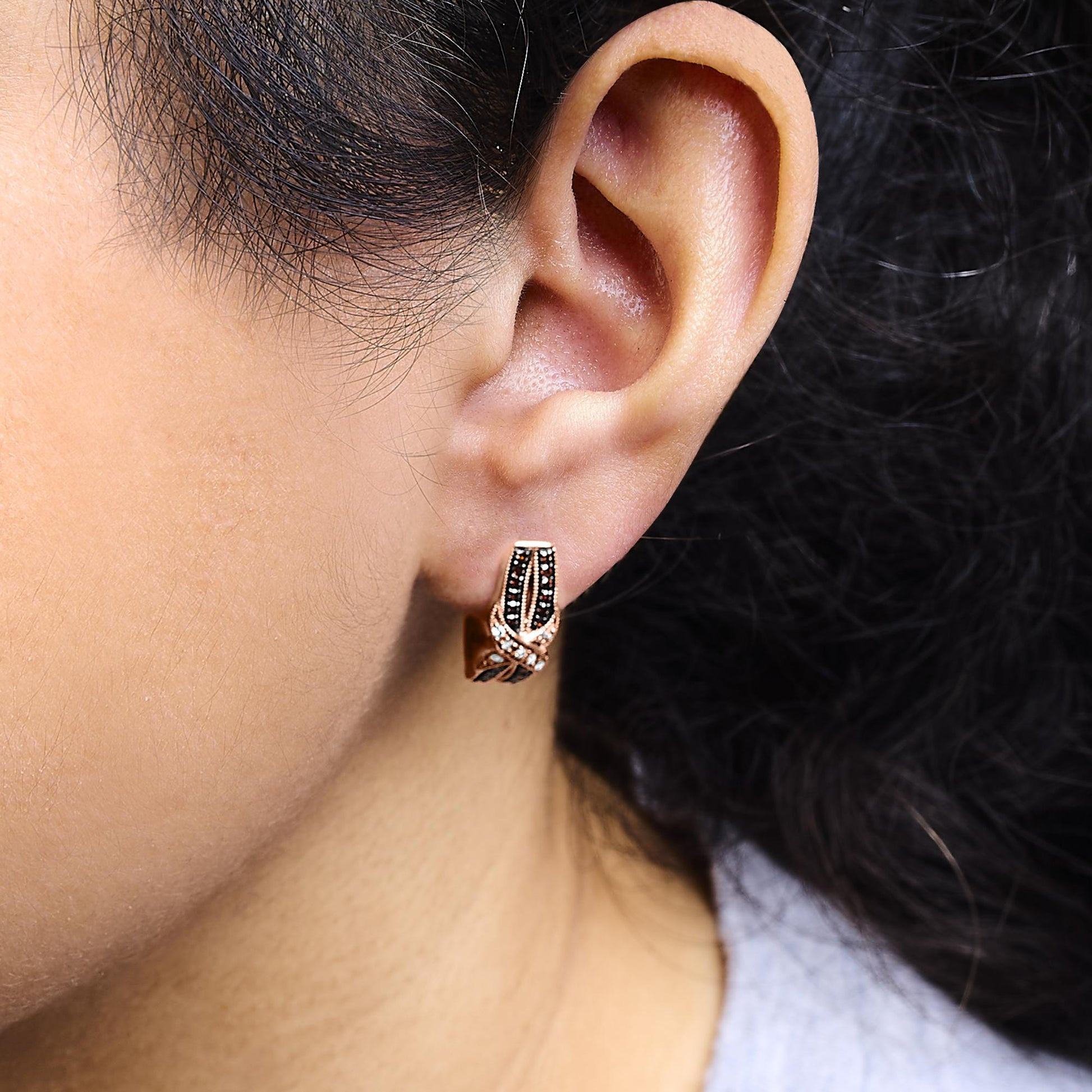 Close-up side view of a ear with a rose-colored metal earring featuring intertwined sections set with small white and brown round gemstones in a pave setting, worn on a model.