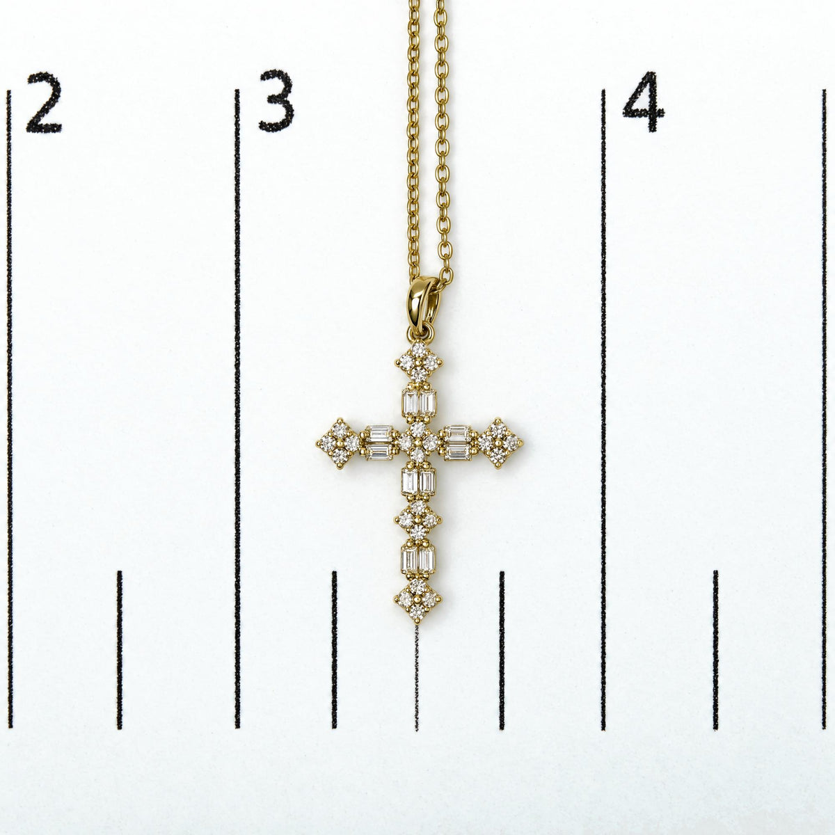 Close-up front view of a yellow metal cross-shaped necklace pendant with round and baguette white gemstones in prong settings, shown on a scale.