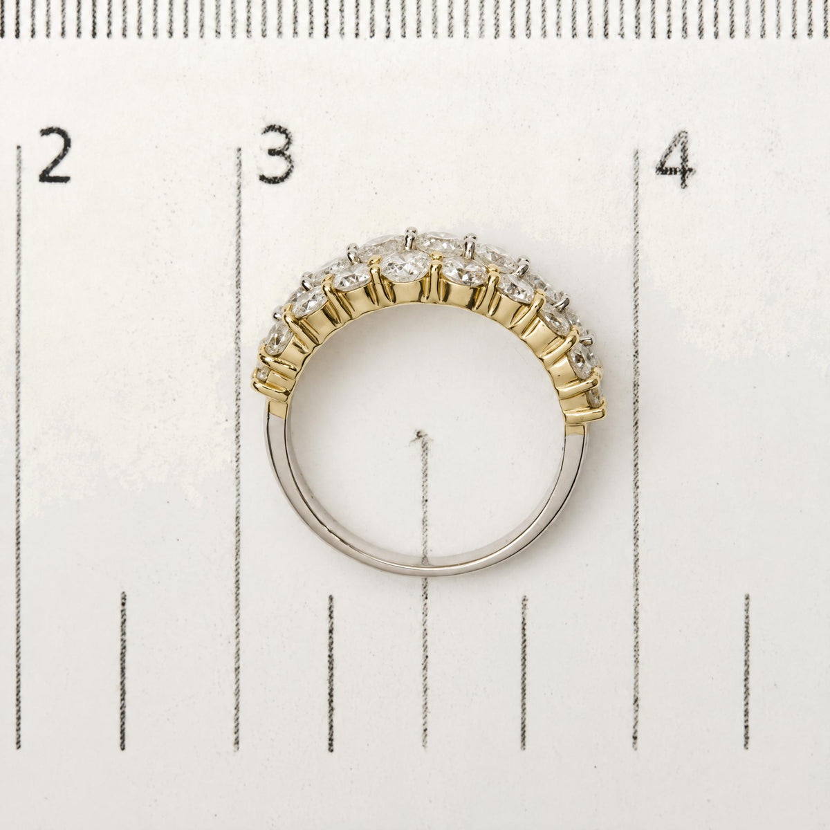 Close-up of a two-tone ring with alternating yellow and white metal bands, set with round white gemstones in prong settings, shown from a top-down angle on a measuring scale.
