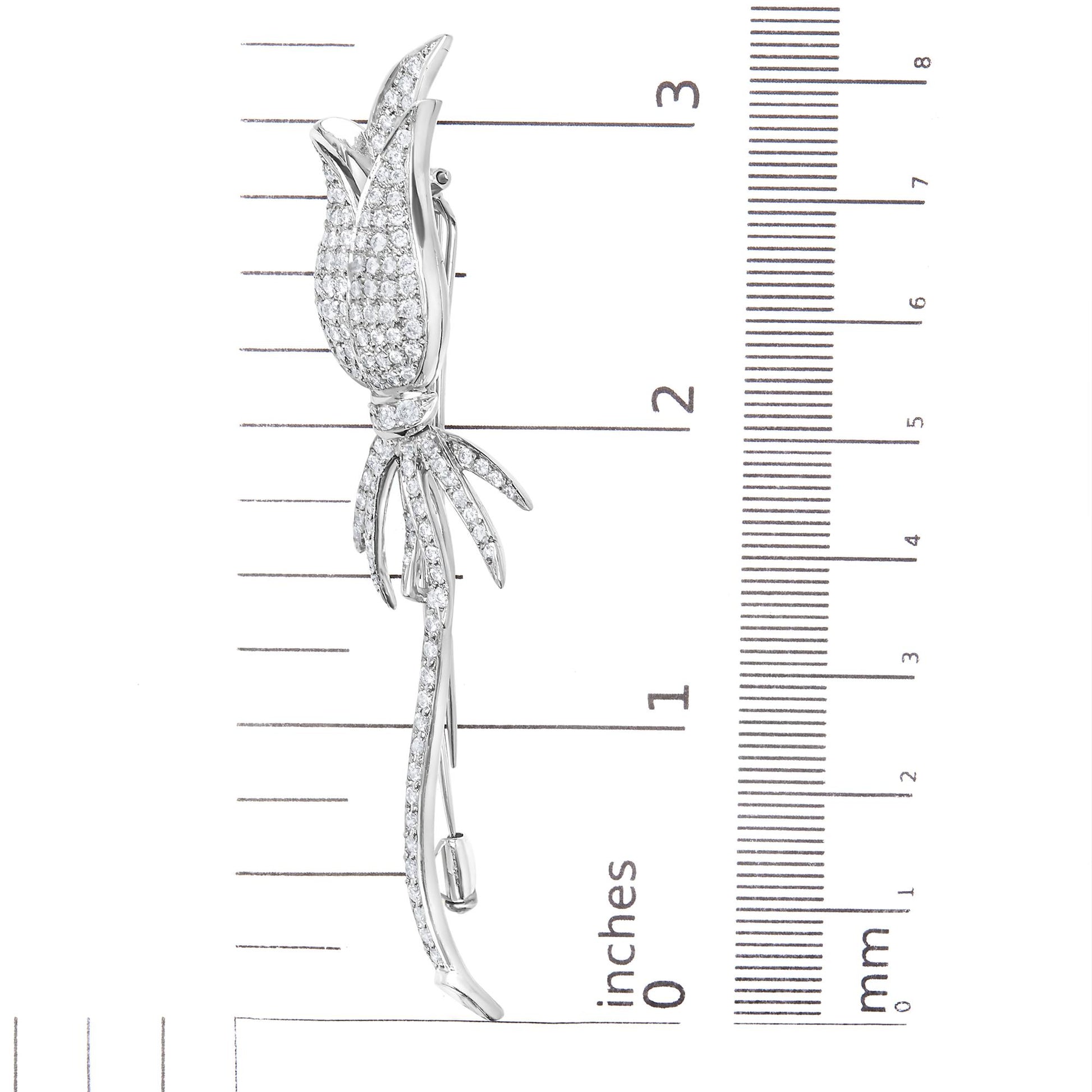 Side view of a white metal flower-shaped brooch with pave-set round white gemstones, placed on a ruler for size reference.