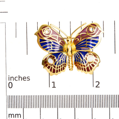 Top-down view of a yellow metal butterfly brooch with blue, red, and white enamel detailing, placed on a ruler for size reference.