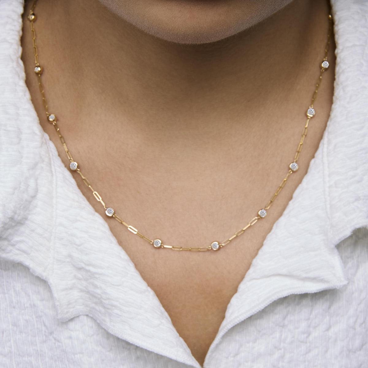 Close-up front view of a model wearing a yellow metal necklace with round white gemstones in bezel settings evenly spaced along a chain.