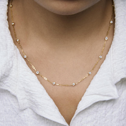 Close-up front view of a model wearing a yellow metal necklace with round white gemstones in bezel settings evenly spaced along a chain.