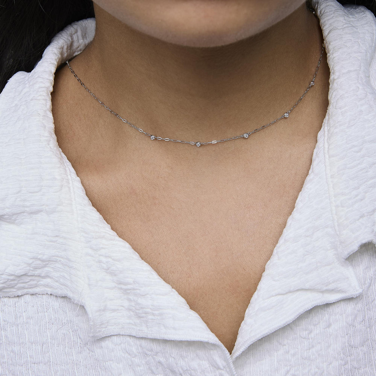 Close-up front view of a model wearing a delicate white necklace with round bezel-set white gemstones evenly spaced along the chain.