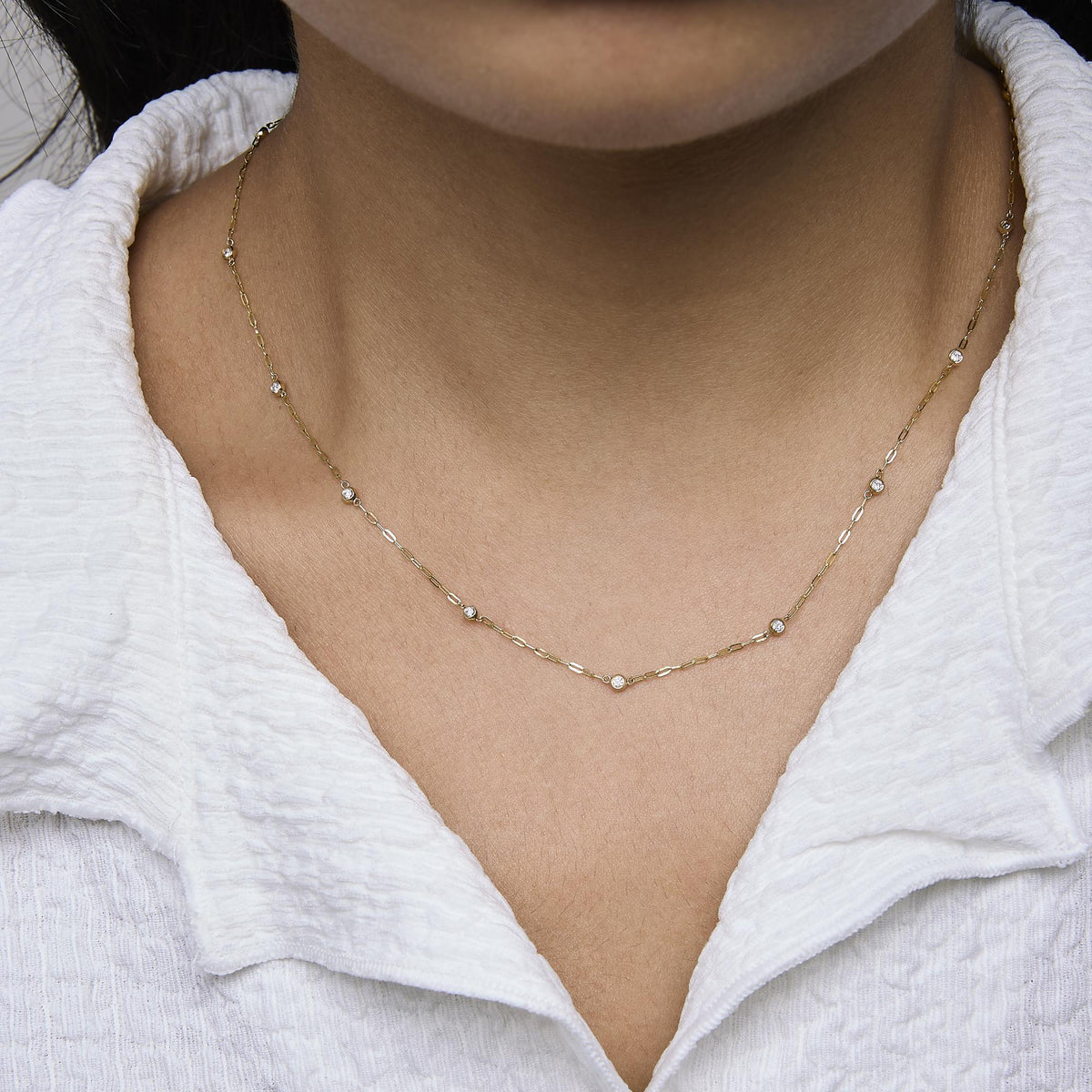 Close-up front view of a model wearing a yellow metal necklace with evenly spaced round white gemstones in bezel settings along a delicate chain.