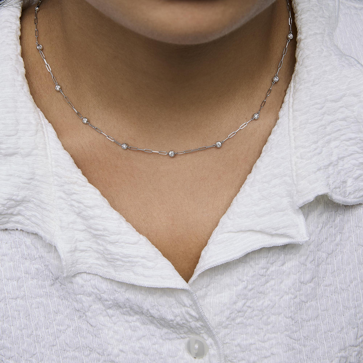 Close-up front view of a model wearing a white metal necklace with round white gemstones in bezel settings evenly spaced along a chain with elongated links.