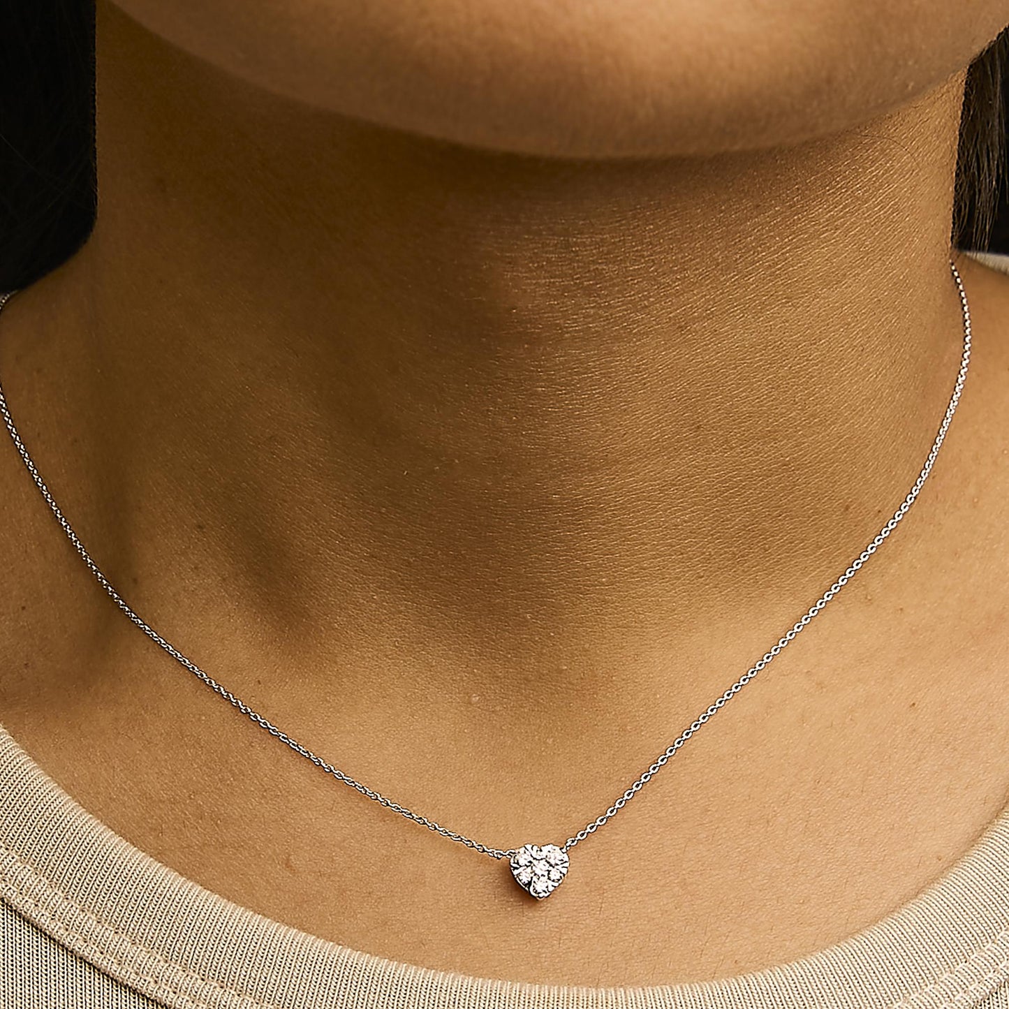 Close-up of a model wearing a white metal necklace with a heart-shaped pendant featuring seven round white gemstones in an invisible setting, shot from the front.