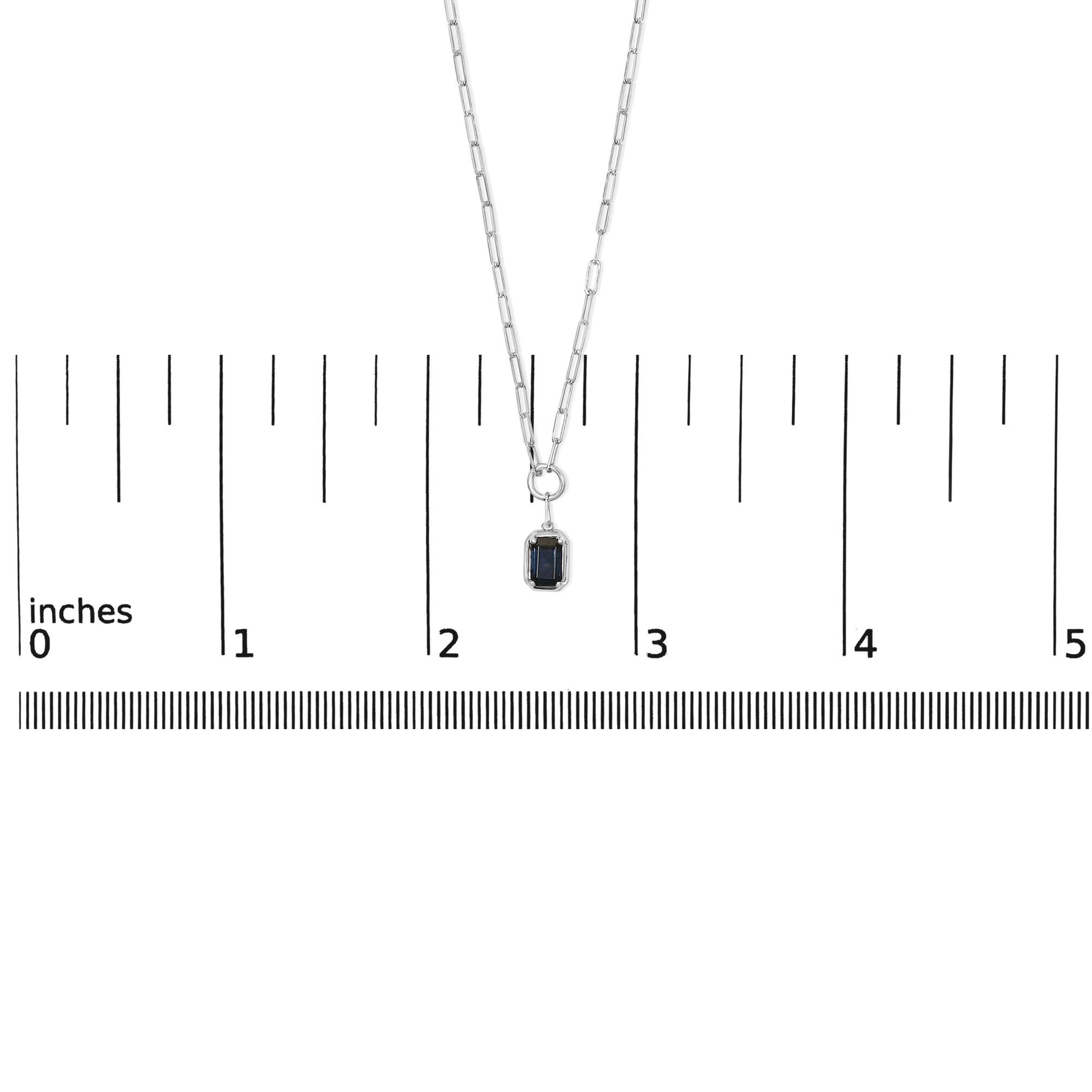 White metal necklace with a blue emerald-shaped gemstone pendant in bezel setting, shown on a ruler scale, front view.
