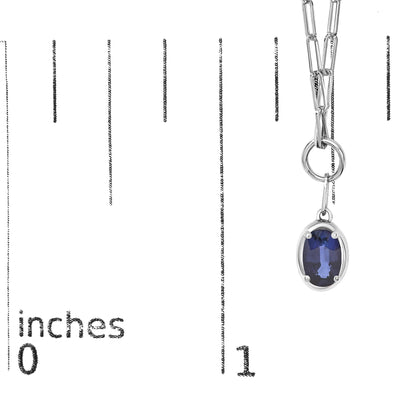Close-up front angle of a white metal necklace with an oval-shaped blue gemstone pendant in a prong setting, shown on a measurement scale.