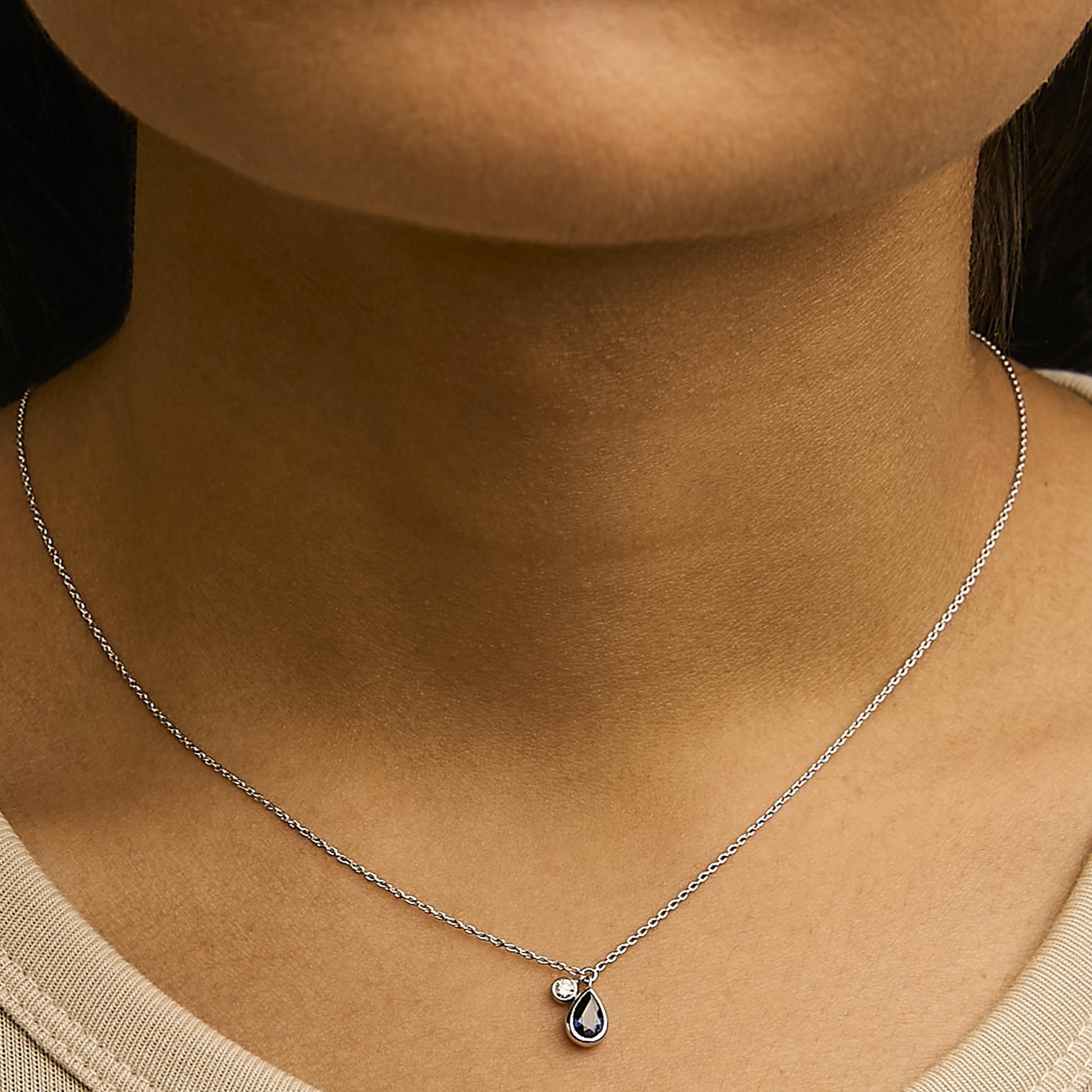 Close-up front view of a model wearing a white metal necklace with a round white gemstone and a pear-shaped blue gemstone in bezel settings.