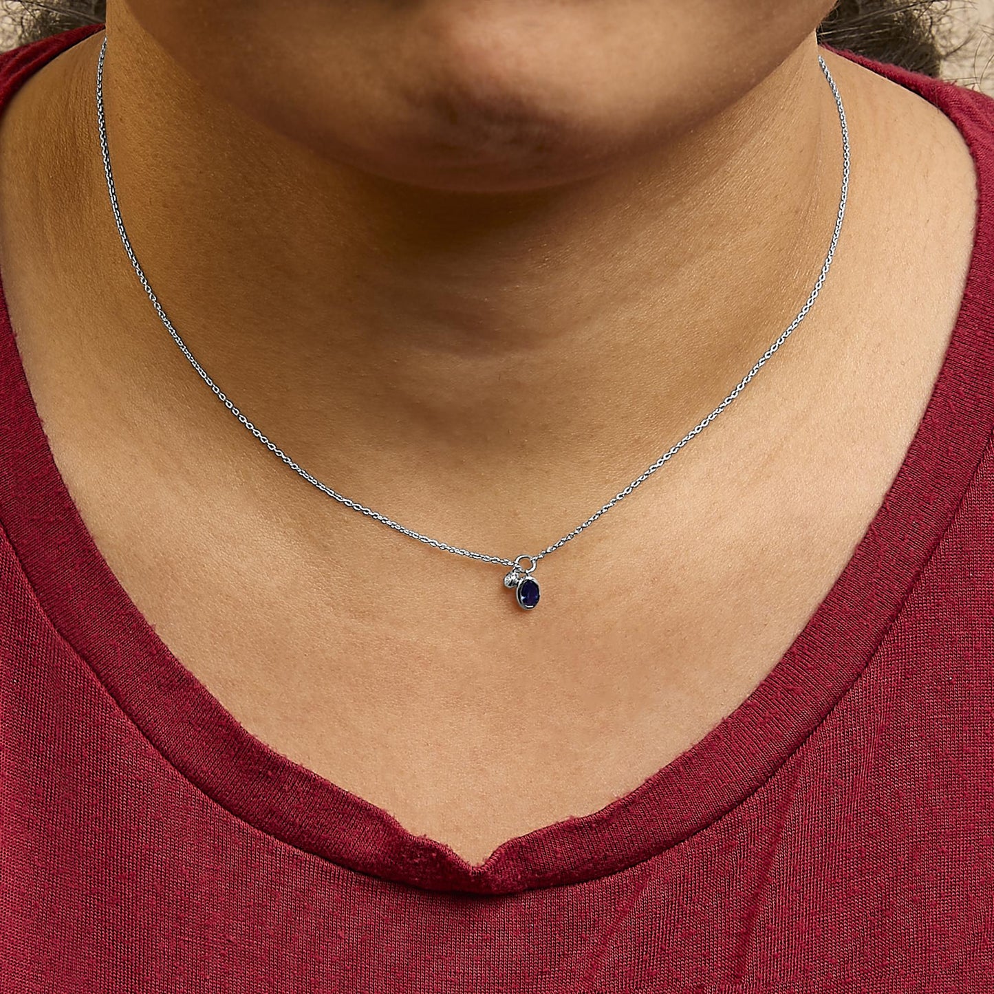 Close-up front view of a white metal necklace worn on a model's neck, featuring a small oval blue gemstone and a round white gemstone, both in bezel settings, hanging from a delicate chain.