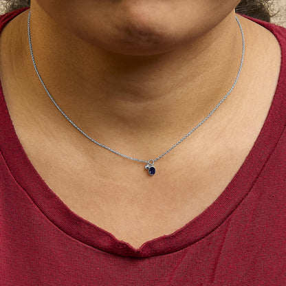 Close-up front view of a white metal necklace worn on a model's neck, featuring a small oval blue gemstone and a round white gemstone, both in bezel settings, hanging from a delicate chain.