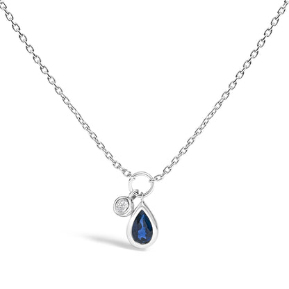 White metal necklace with a chain and two charms, shown in a front-facing close-up. One charm features a pear-shaped blue gemstone in a bezel setting, and the other has a round white gemstone in a bezel setting.