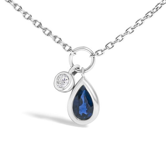 Close-up front angle of a white metal chain necklace with two bezel-set pendants: a round white gemstone and a pear-shaped blue gemstone.