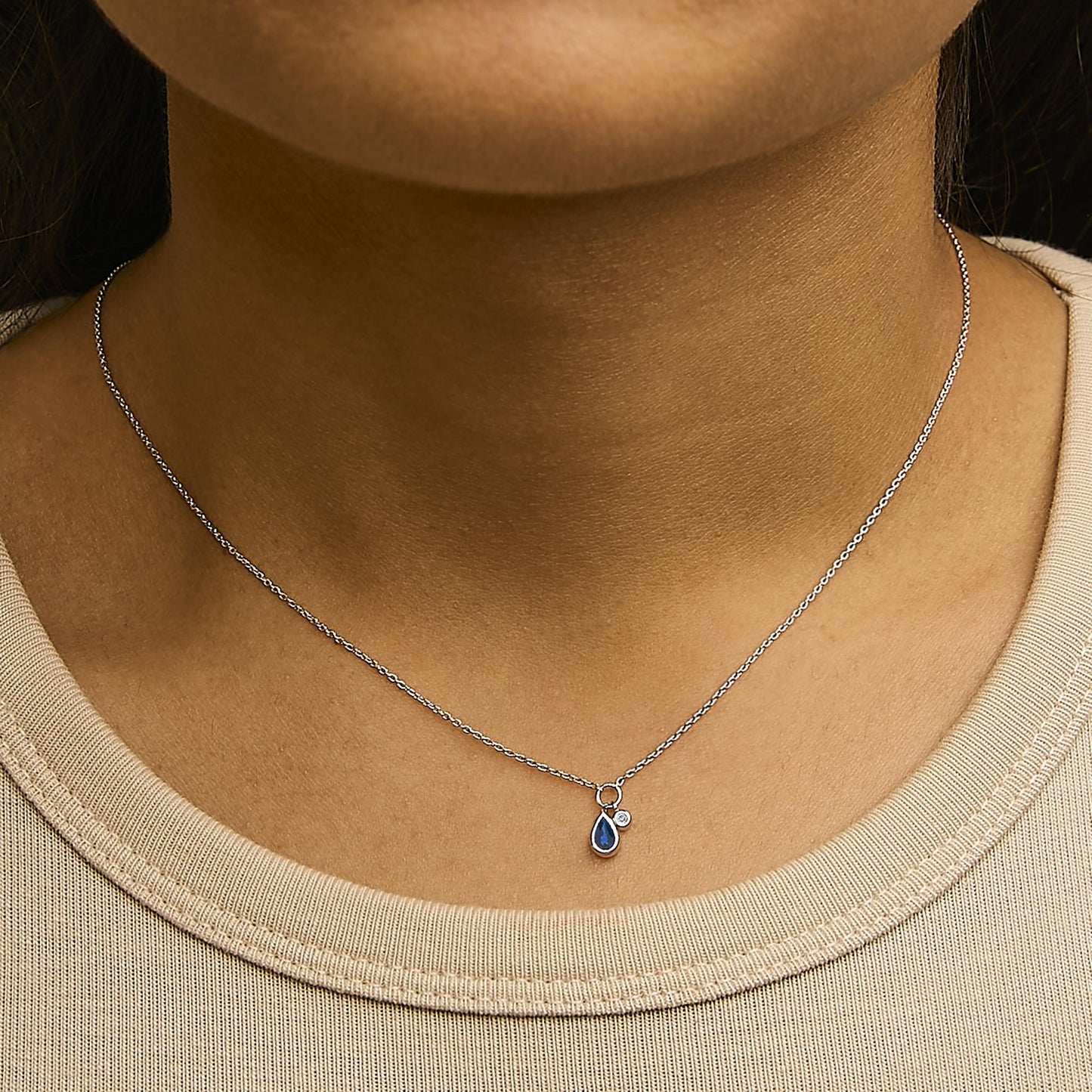 Close-up front view of a model wearing a white metal necklace with a pear-shaped blue gemstone and a small round white gemstone pendant, both in bezel settings.