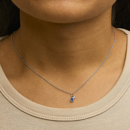 Close-up front view of a model wearing a white metal necklace with a pear-shaped blue gemstone and a small round white gemstone pendant, both in bezel settings.