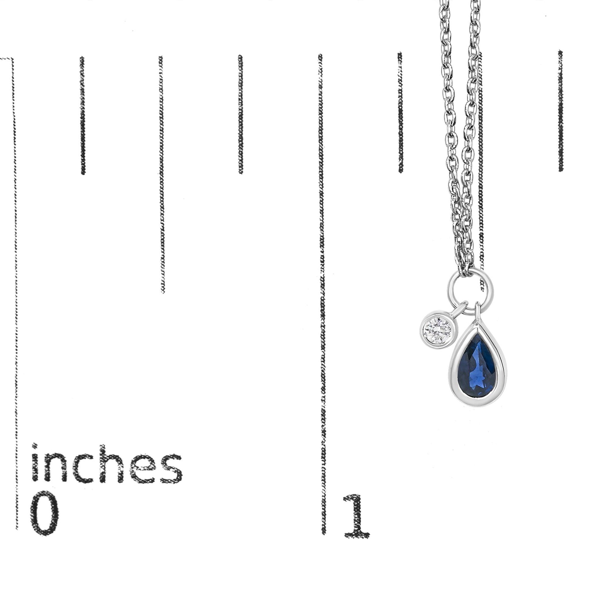 Close-up on a white necklace with a pear-shaped blue gemstone and a round white gemstone, both in bezel settings, shown hanging on a scale.