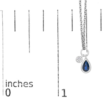 Close-up on a white necklace with a pear-shaped blue gemstone and a round white gemstone, both in bezel settings, shown hanging on a scale.