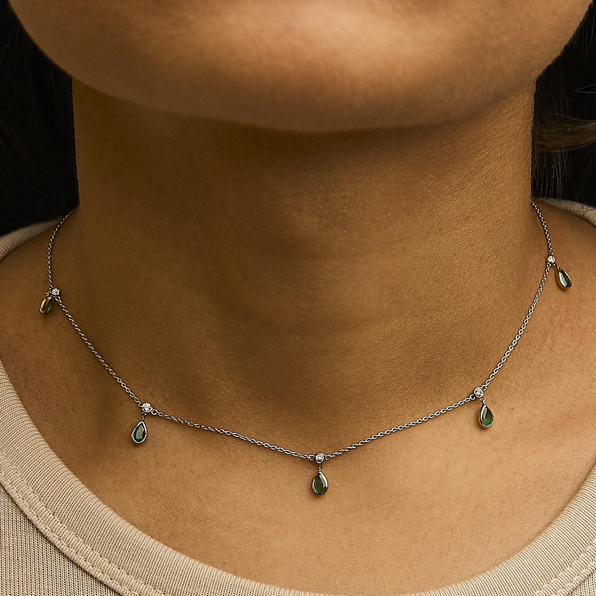 Necklace with pear-shaped green gemstones and round white gemstones in bezel settings on a yellow metal chain, shown worn on a model, front close-up angle.