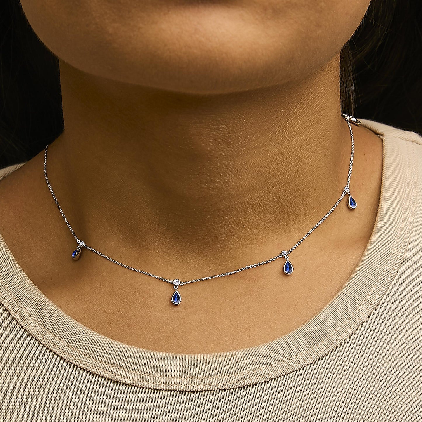 White necklace with a delicate chain featuring pear-shaped blue gemstones and round white gemstones in bezel settings, shown worn on a model's neck from a front angle.