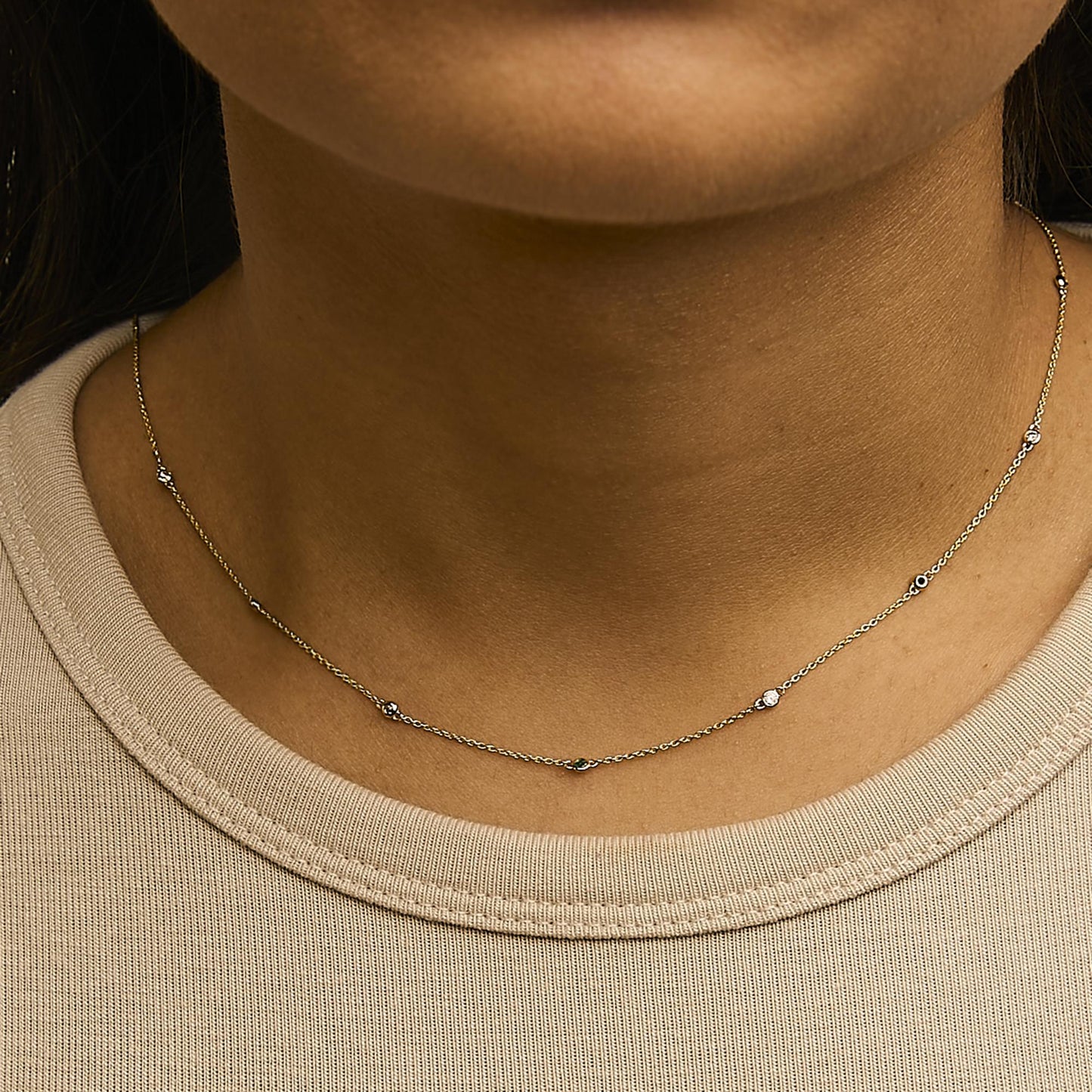 Close-up front view of a yellow metal necklace worn on a model's neck, featuring a delicate chain with alternating small round white gemstones and round green gemstones in bezel settings.