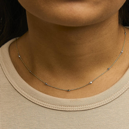 Close-up front view of a yellow metal necklace worn on a model's neck, featuring a delicate chain with alternating small round white gemstones and round green gemstones in bezel settings.