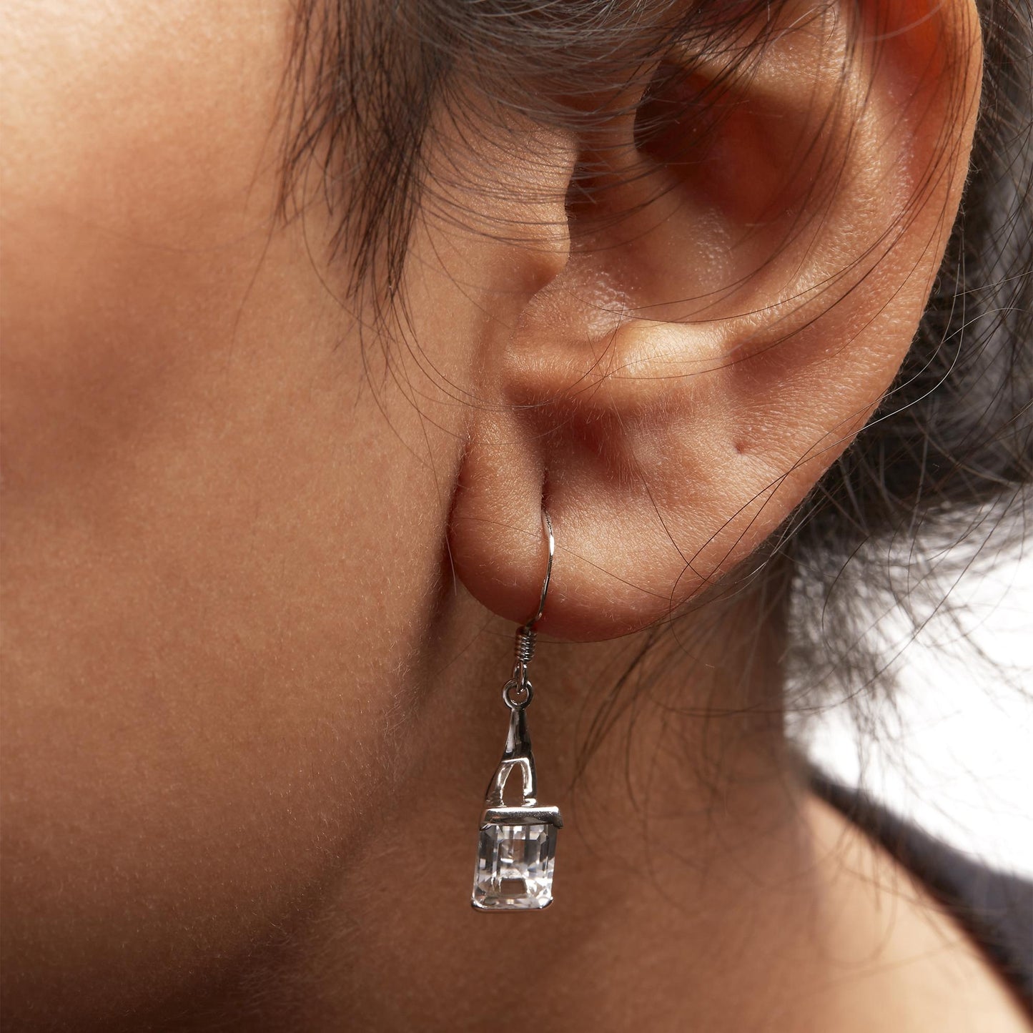 Close-up side view of a white metal dangle earring with a white emerald-shaped gemstone in a 4-prong setting worn on a model.