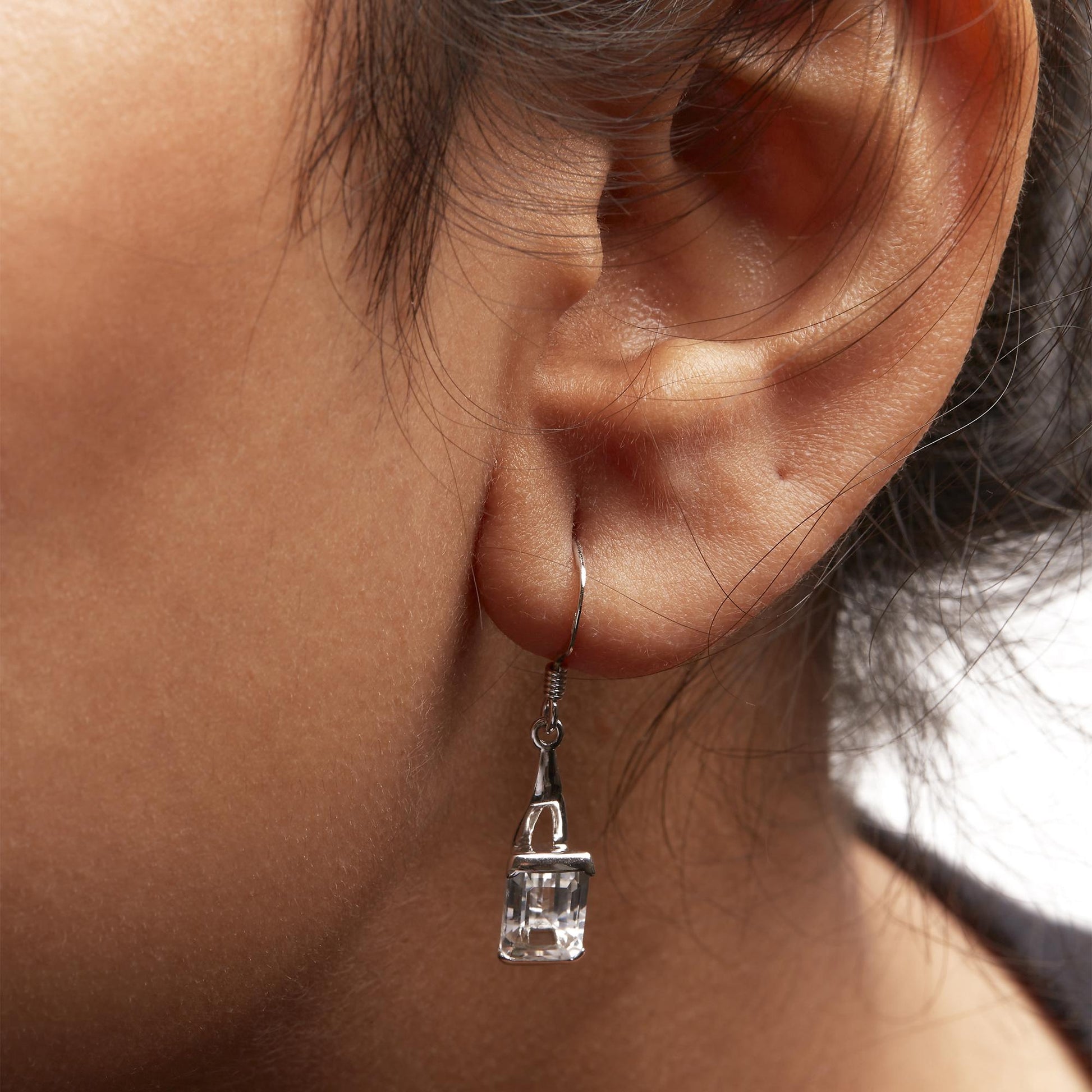 Close-up side view of a white metal dangle earring with a white emerald-shaped gemstone in a 4-prong setting worn on a model.