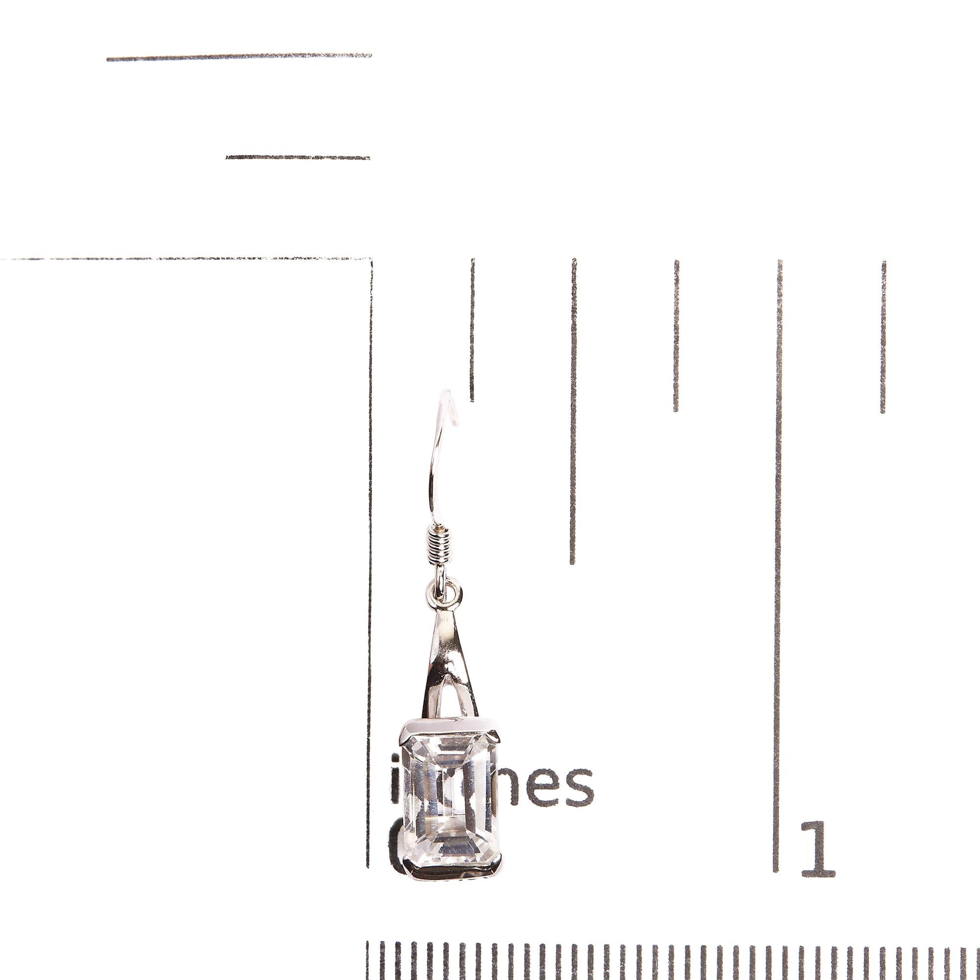 Single white metal earring with a white, emerald-shaped gemstone in a four-prong setting, photographed on a ruler from a front angle.