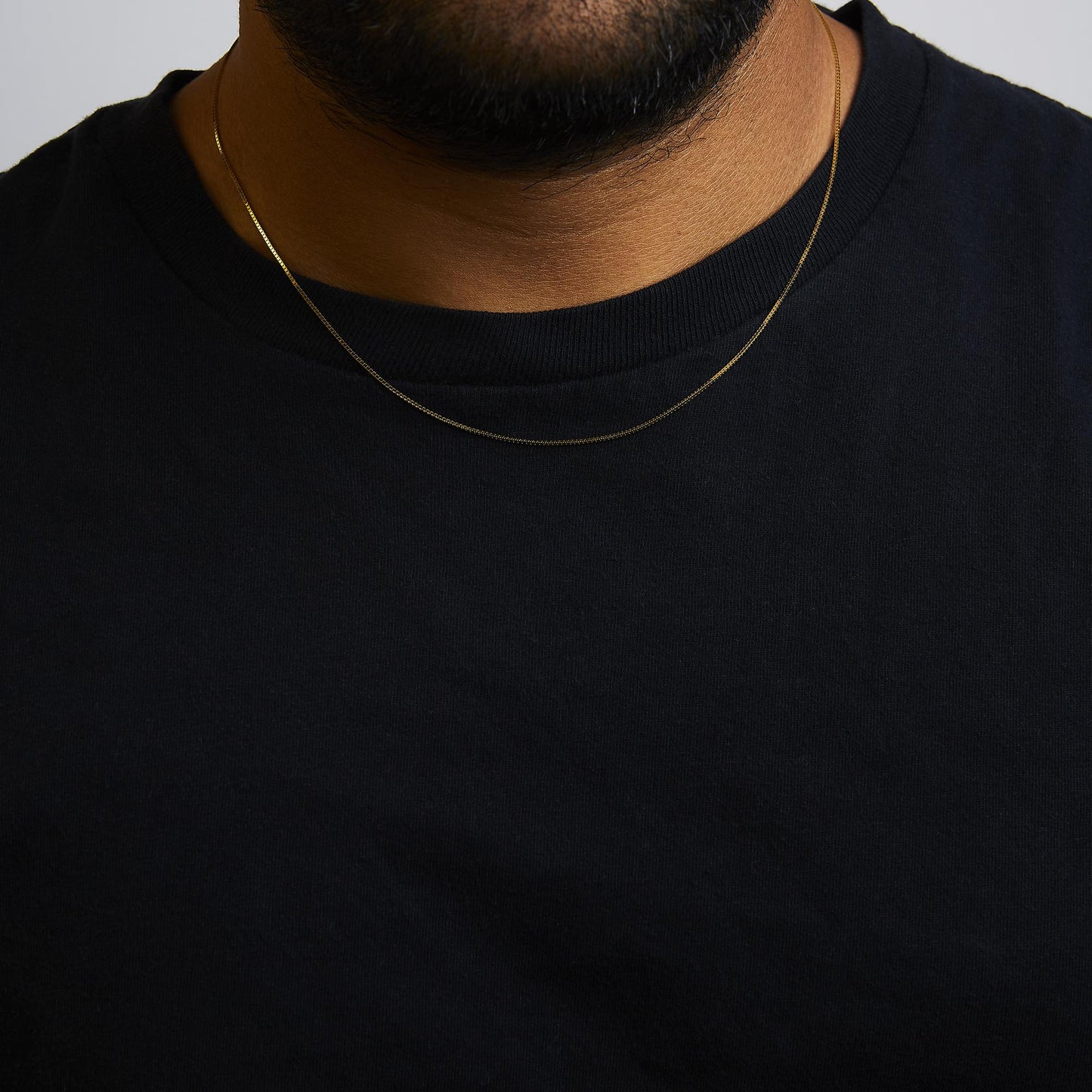 Close-up front view of a thin yellow metal chain necklace worn on a model's neck over a black shirt.