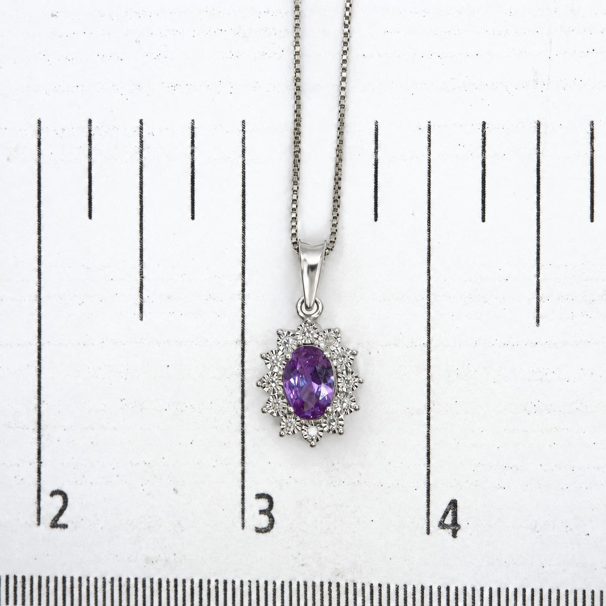 Close-up top-down image of a white metal necklace with a pendant featuring one oval-shaped purple gemstone surrounded by round white gemstones, shown on a measurement scale.