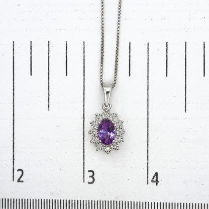 Close-up top-down image of a white metal necklace with a pendant featuring one oval-shaped purple gemstone surrounded by round white gemstones, shown on a measurement scale.