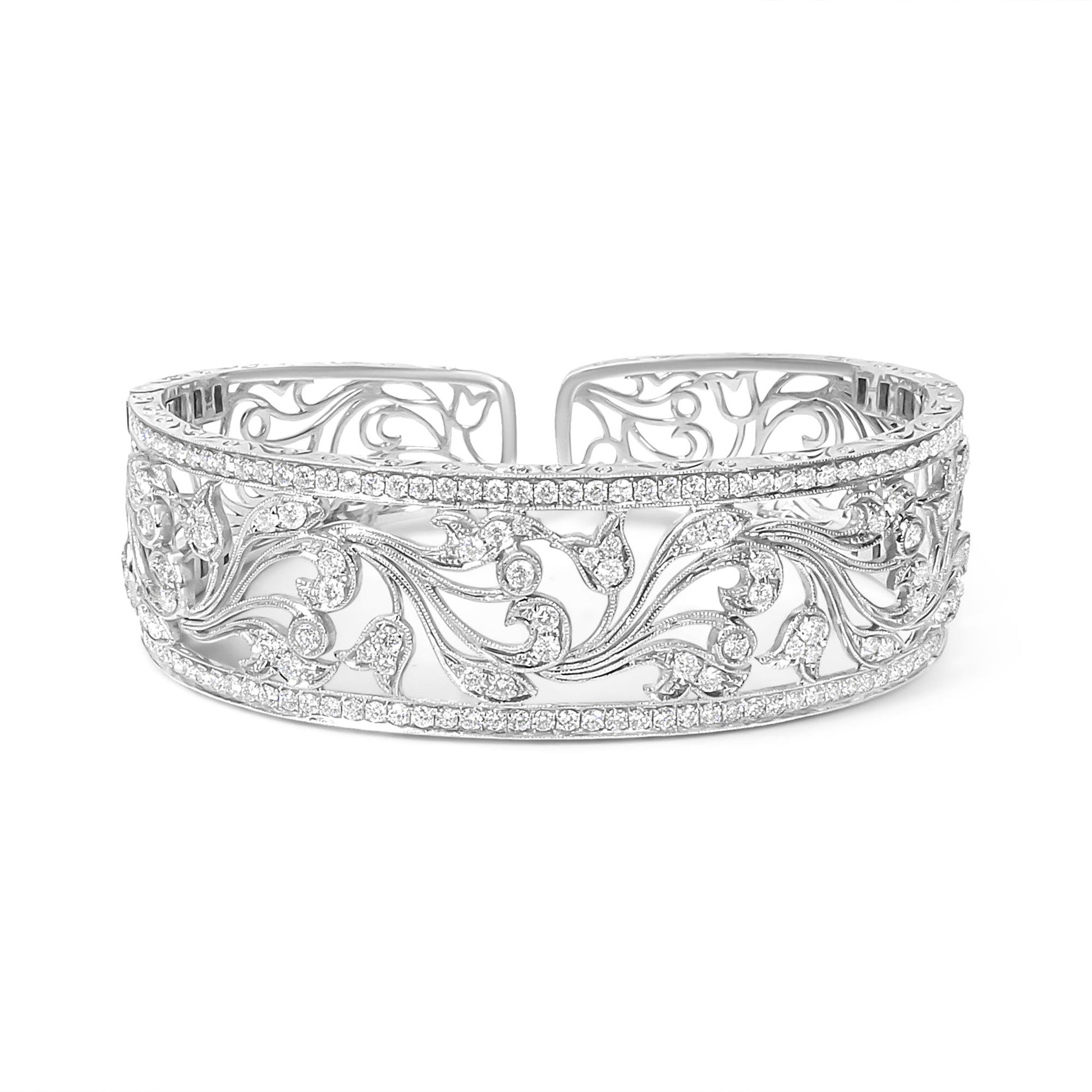 Front-facing shot of an open white metal bracelet with intricate floral scrollwork and rows of round white gemstones in pave setting along the top and bottom edges.