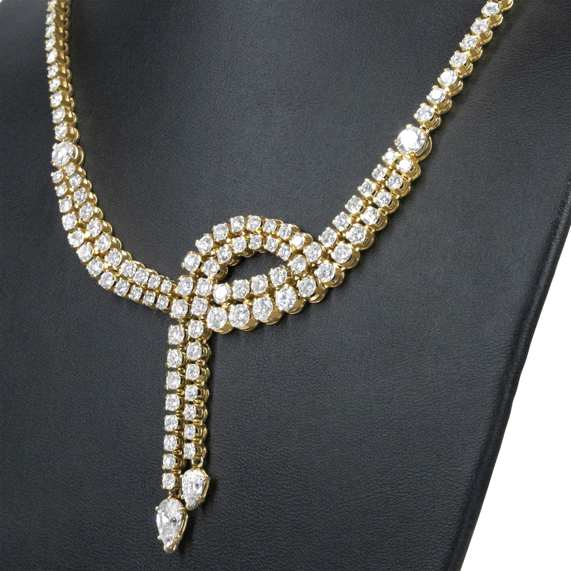 Close-up front angle of a yellow metal necklace with multiple white round gemstones forming a looped design and two dangling strands ending in pear-shaped white gemstones, displayed on a black jewelry stand.