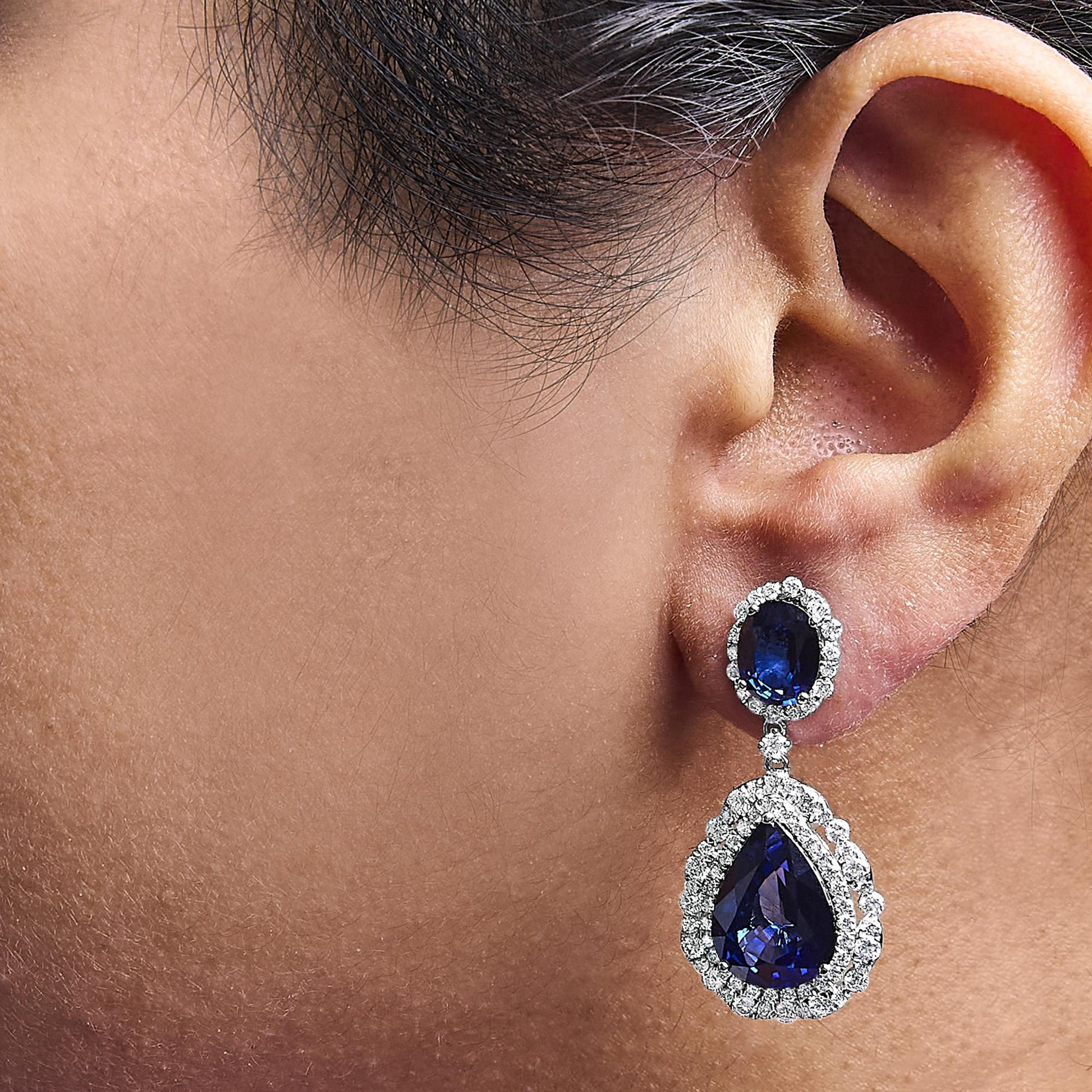 Close-up side view of a white metal drop earring on a model’s ear featuring an oval blue gemstone at the top surrounded by small white gemstones, connected to a larger pear-shaped blue gemstone also surrounded by small white gemstones.