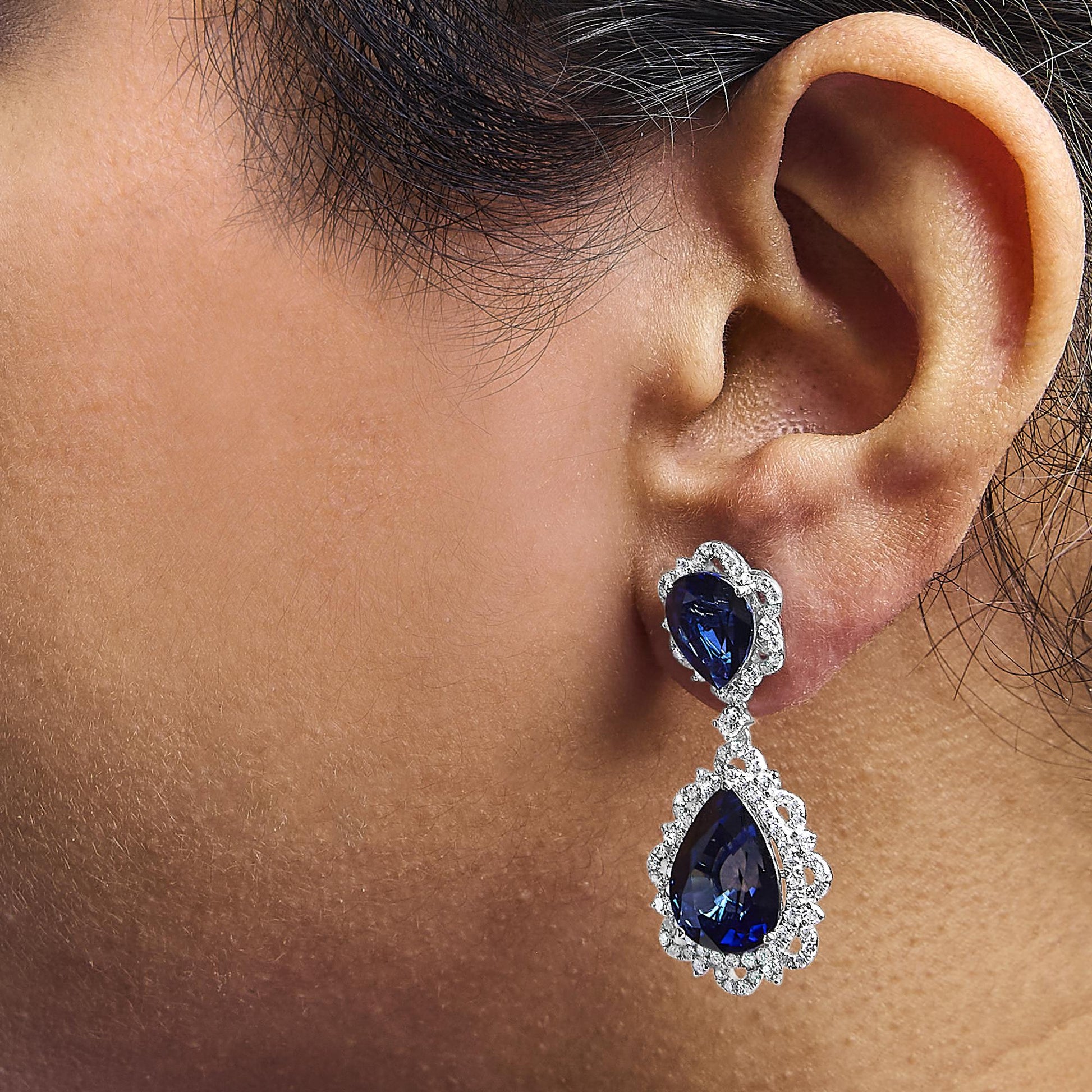 Close-up side view of a white metal drop earring on a model, featuring pear-shaped blue gemstones surrounded by round white gemstones in prong settings.