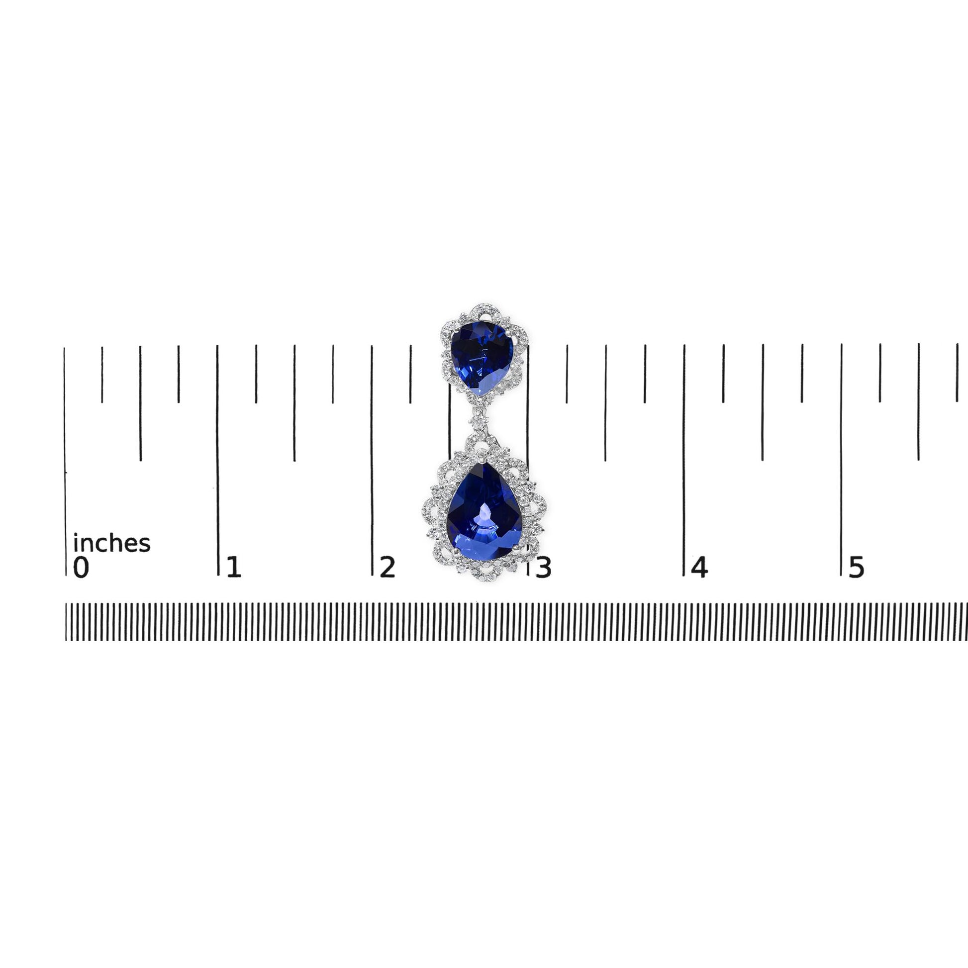 Single white metal earring with two pear-shaped blue gemstones surrounded by white gemstones, shown from above on a ruler for size reference.