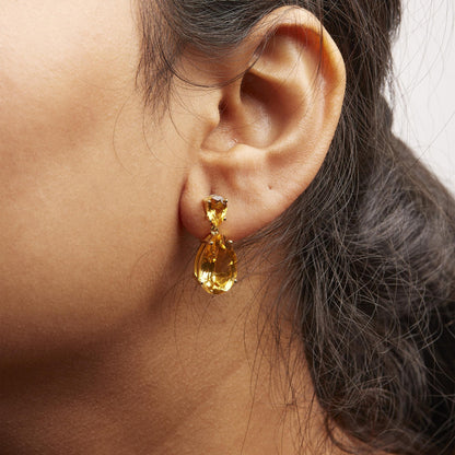 Close-up side view of a model wearing yellow metal earrings with two dangling pear-shaped orange gemstones in four-prong settings.