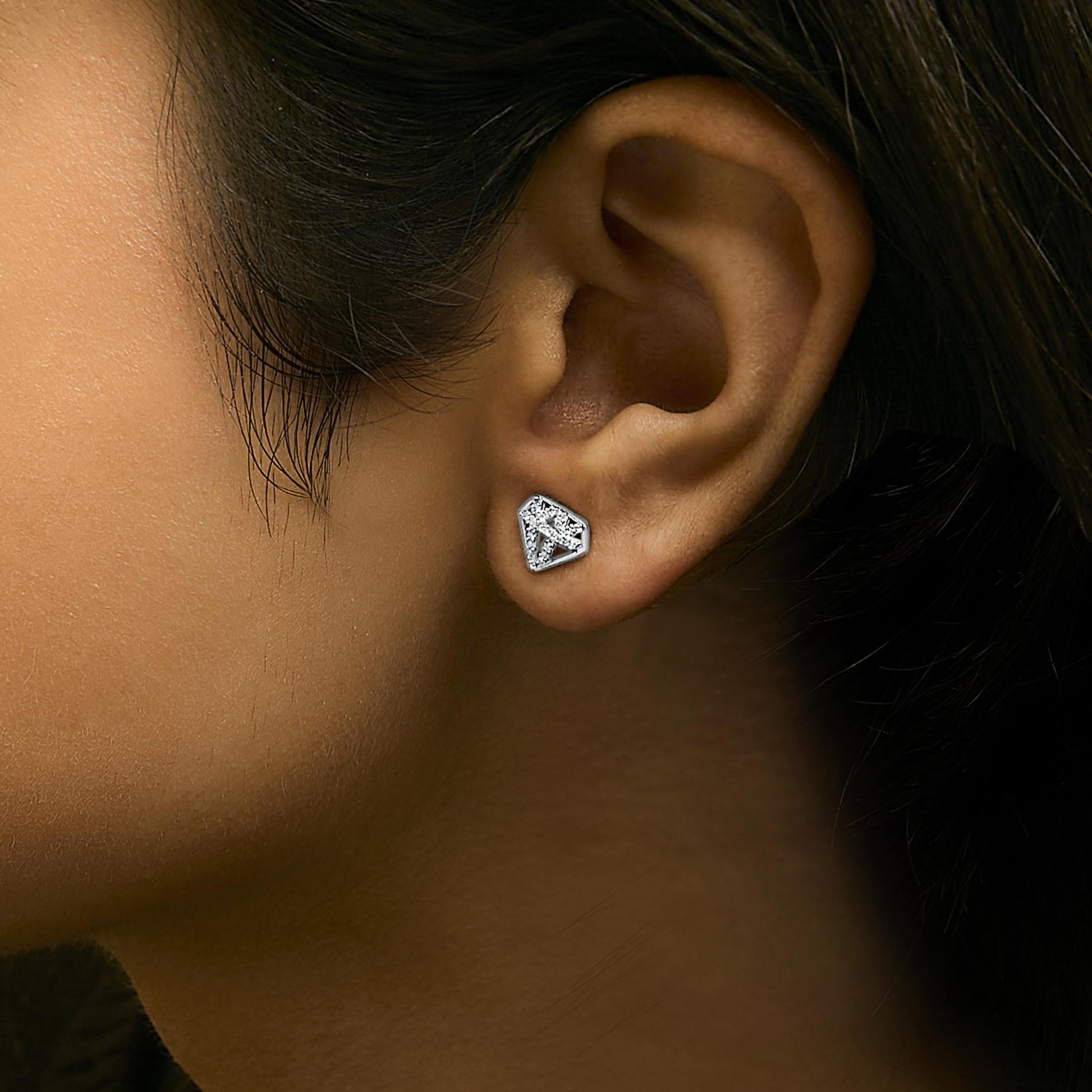 Close-up side view of a model wearing a white metal earring shaped like a geometric pentagon, adorned with round white gemstones in prong settings.