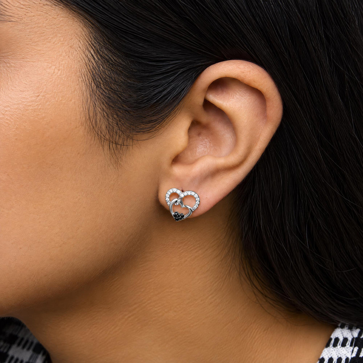 Close-up side view of a model wearing white metal earrings shaped as two interconnected hearts, the larger heart encrusted with white gemstones and the smaller heart featuring black gemstones at the bottom.