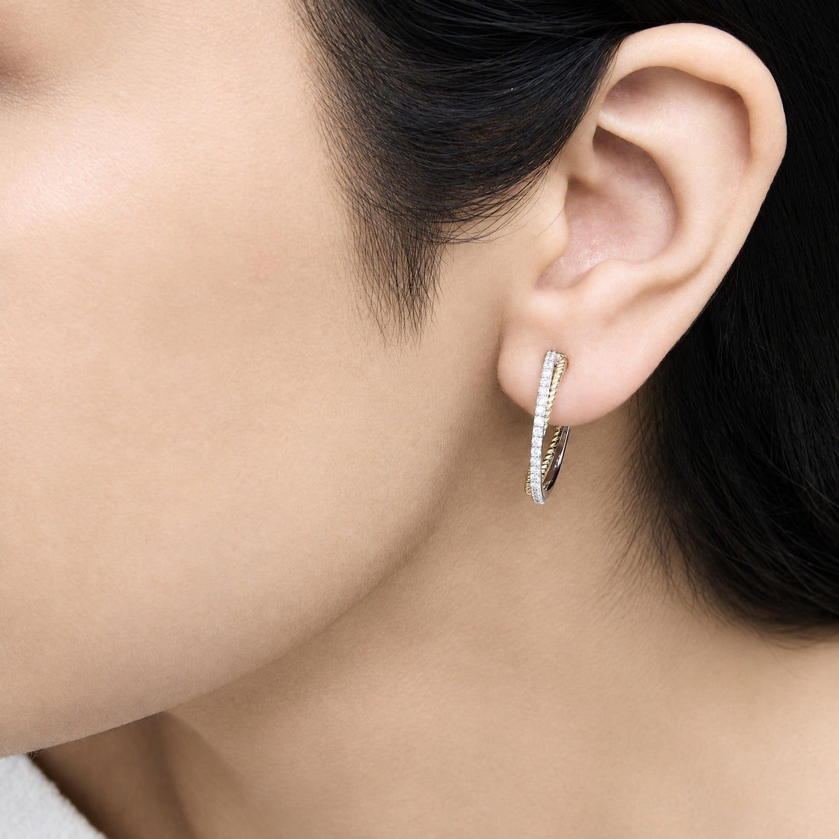 Close-up side view of a model wearing hoop earrings in yellow and white metal with shared prong set round white gemstones.