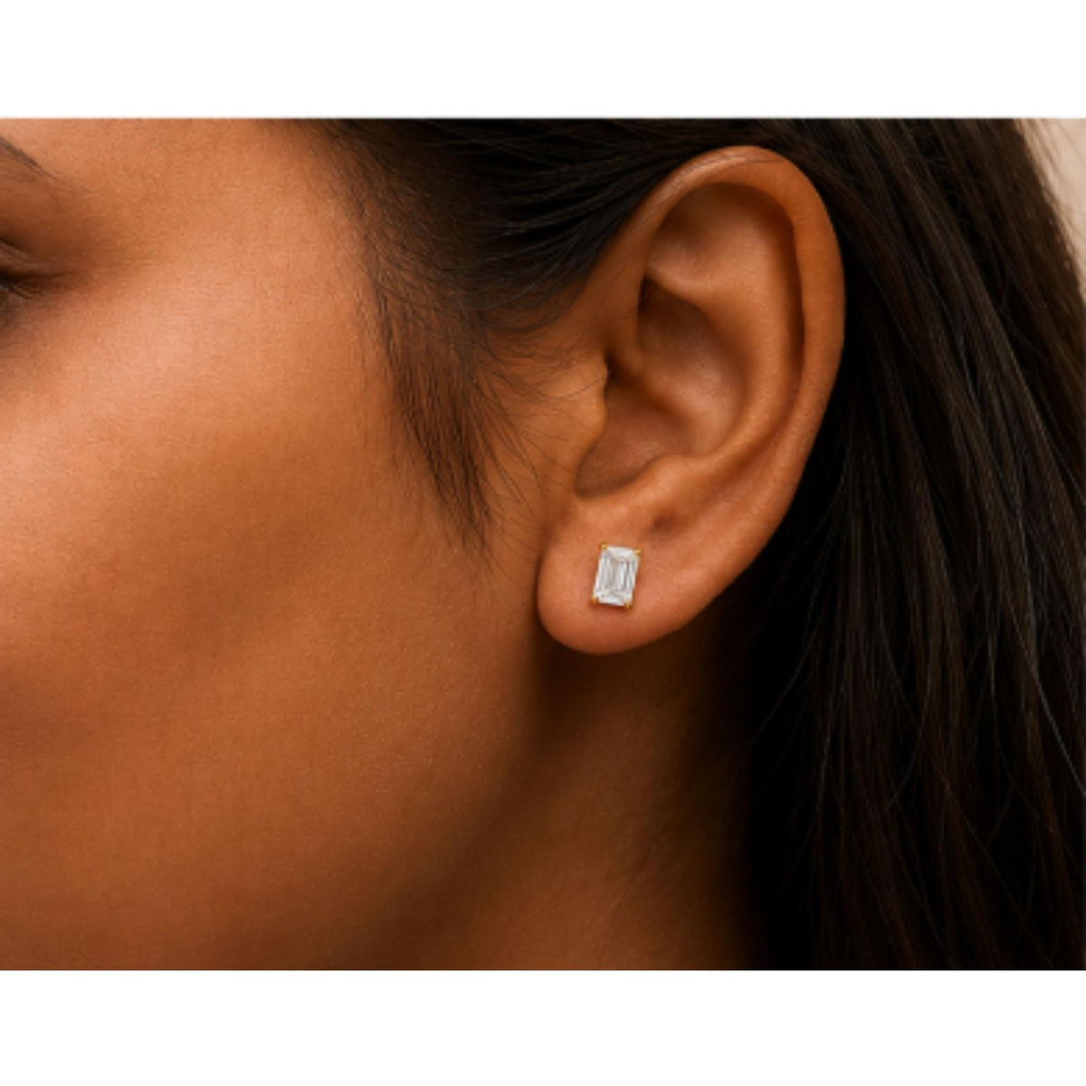 Close-up side view of a model wearing a pair of yellow metal stud earrings with white emerald-shaped gemstones in a 4-prong setting.