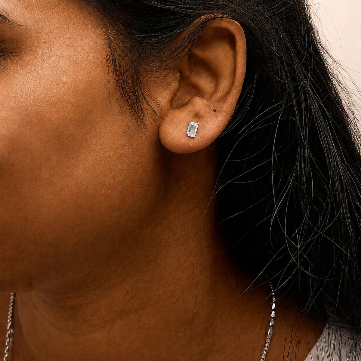 Close-up side view of a model wearing a white metal stud earring with a rectangular emerald-shaped white gemstone in a four-prong setting.