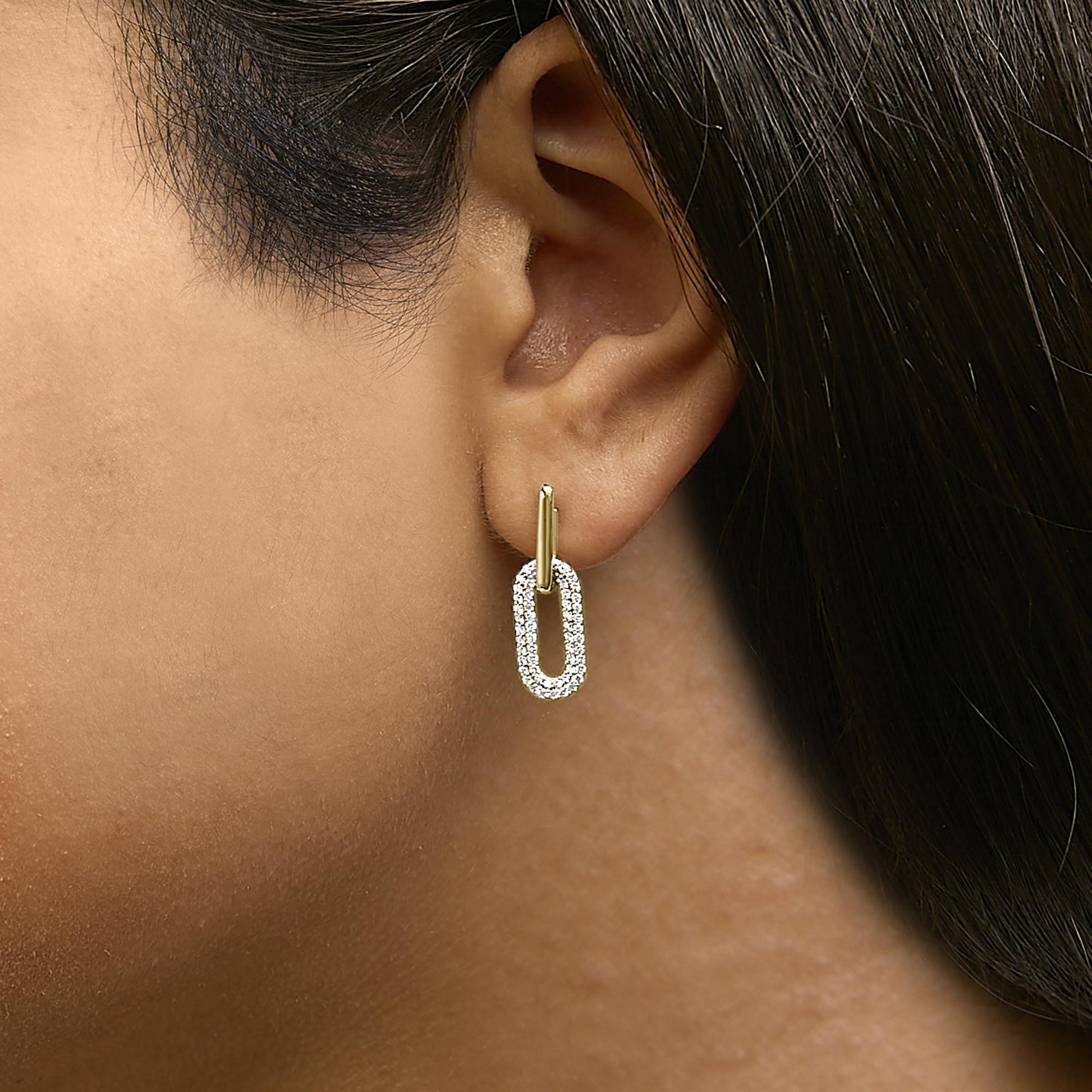 Close-up side view of a model wearing yellow metal earrings with a vertical bar connecting to a hanging oval hoop encrusted with white round gemstones in shared prong settings.