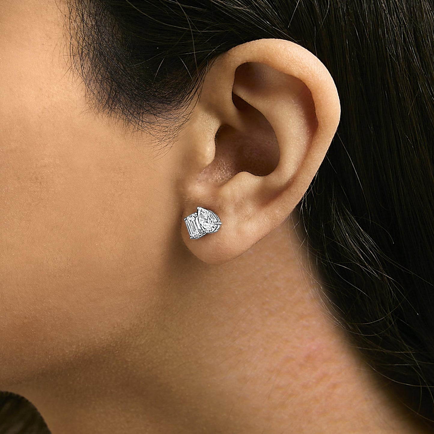 Close-up side view of a white metal earring worn on a model’s ear, featuring one emerald-shaped white gemstone in a four-prong setting and one pear-shaped white gemstone in a three-prong setting.