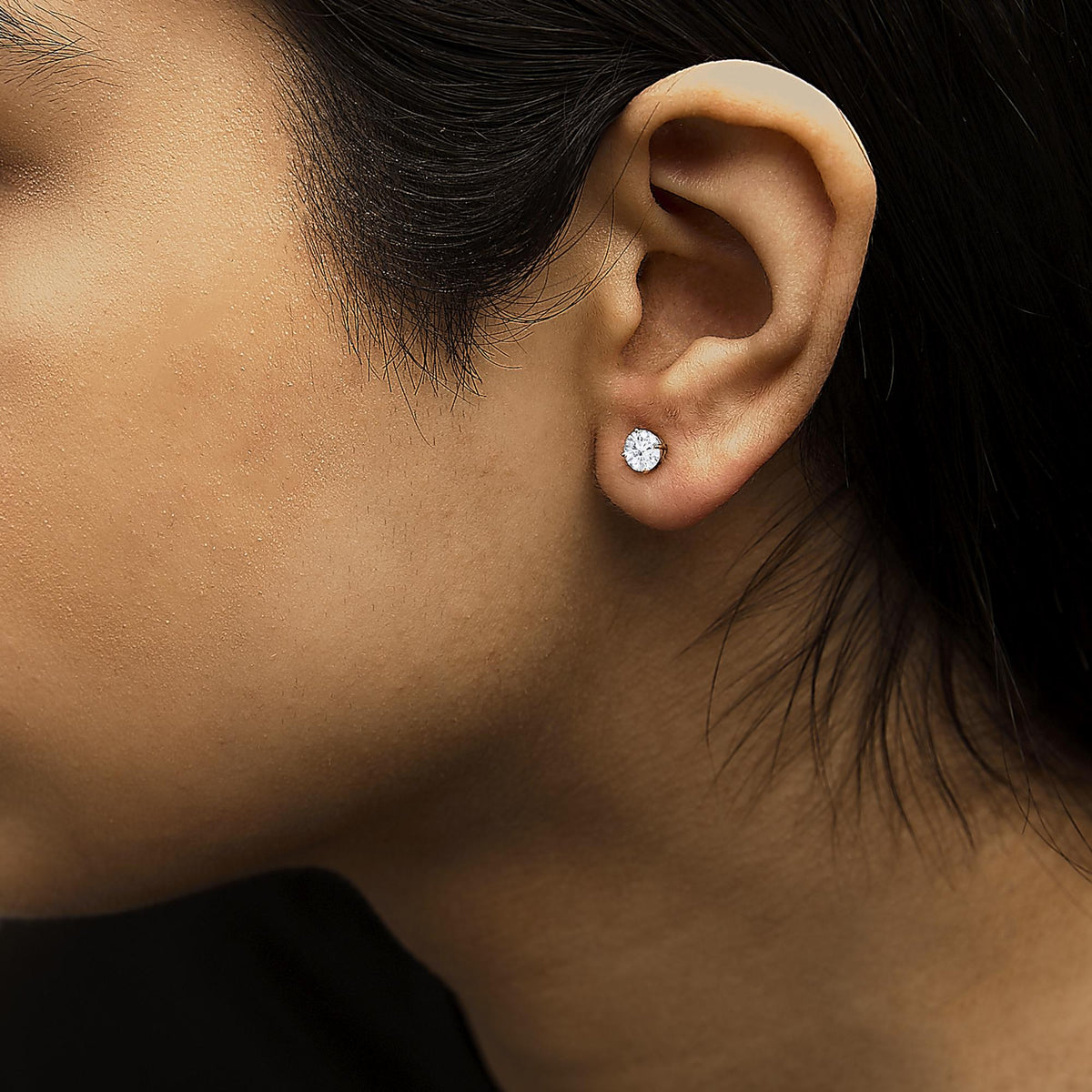 Close-up side view of a model wearing a yellow metal stud earring with a round white gemstone in a four-prong setting.