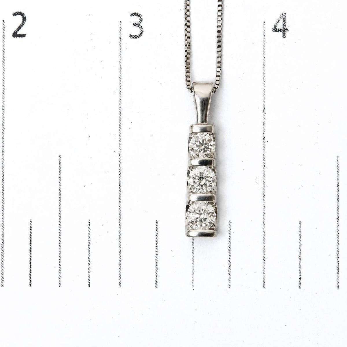 Close-up image of a white metal necklace pendant with three round white gemstones in a channel setting, shown on a ruler scale with measurements visible, shot from the front.