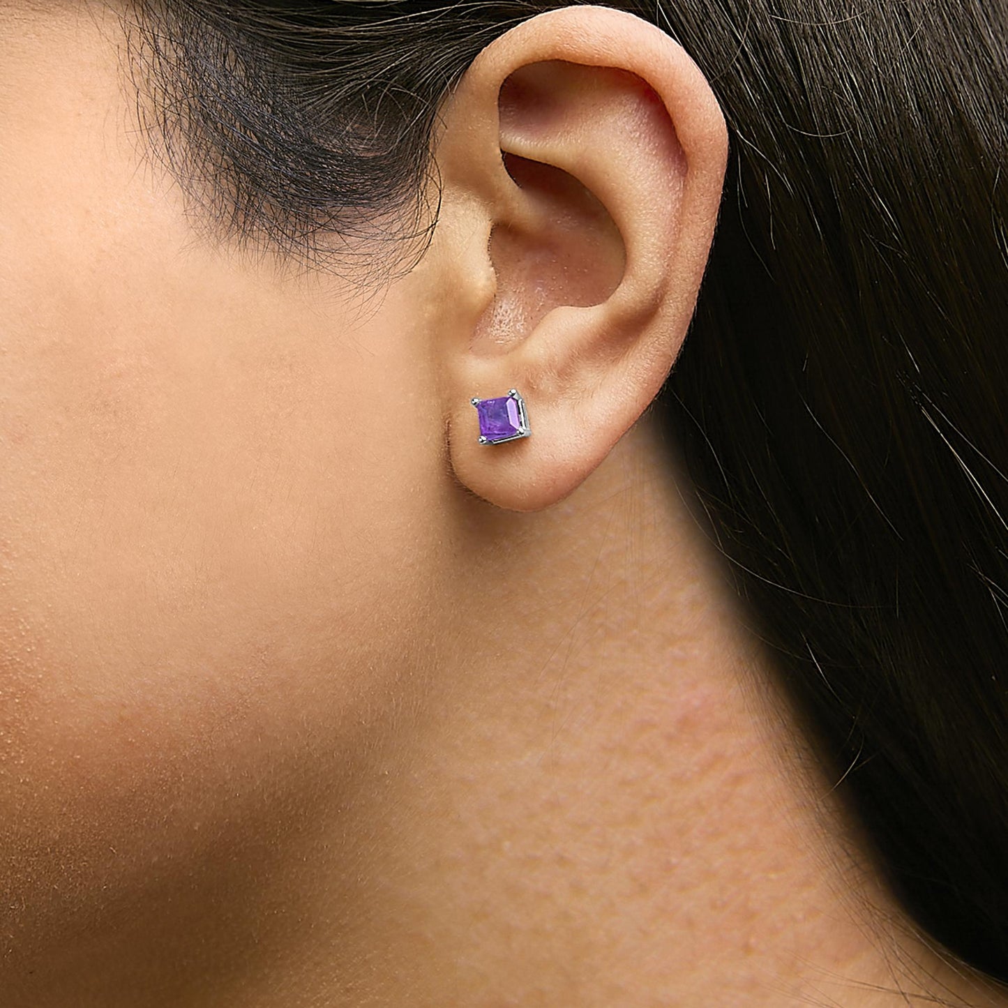 Close-up side angle of a model wearing white metal stud earrings with square purple gemstones in four-prong settings.